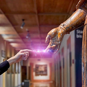 Linke Hand eines Besuchers zeigt mit ausgestrecktem Zeigefinger auf die aus Holz geschnitzte rechte Hand einer Heiligenfigur; zwischen den Fingerspitzen ein violett leuchtender Funken, im Hintergrund unscharfer Museumsflur