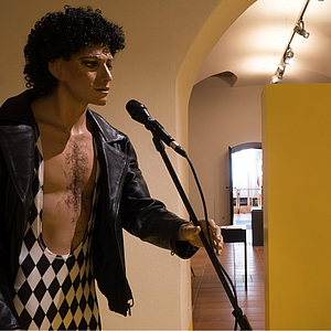 Museumspuppe eines männlichen Sängers mit lockigem Haar, freier Brust und schwarzer Lederjacke, in schwarz-weiß kariertem Outfit am Mikrofon in einem Gewölbegang des Franziskanermuseums.