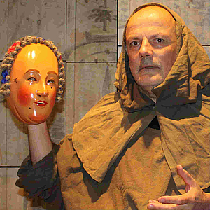 Mann in braunem Franziskanerhabit hält eine bemalte, antik wirkende Holzmaske mit Perücke in der Hand; er blickt ernst vor schlichter Wand