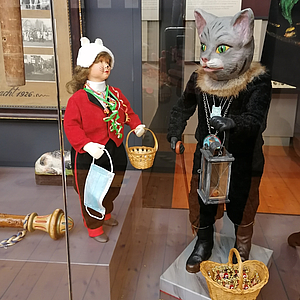 Zwei Ausstellungsfiguren: links ein Kind in roter Jacke, weißer Mütze, mit Korb und OP‑Maske; rechts eine aufrechte Katzenfigur in dunklem Gewand mit Pelzkragen, Laterne und einem Korb voller Süßigkeiten.