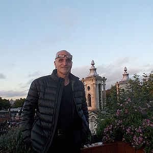Älterer Mann mit Brille auf der Stirn und dunkler Steppjacke steht auf einer Dachterrasse, lächelnd; im Hintergrund zwei barocke Kirchtürme und blühende Pflanzen am Geländer unter hellem Himmel.