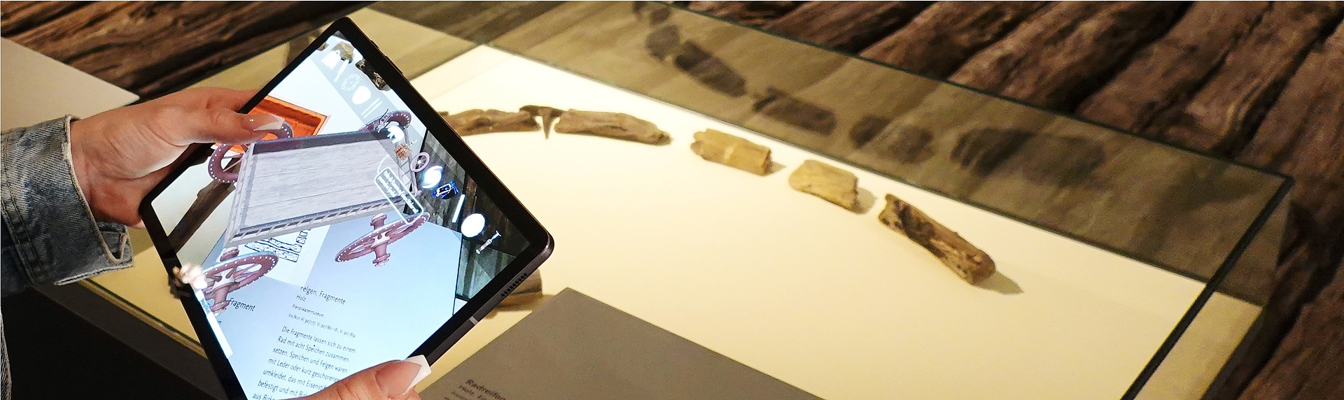 Hände halten ein Tablet mit Museumsinformationen; daneben eine Glasvitrine mit mehreren kleinen archäologischen Fragmenten auf weißer Unterlage.