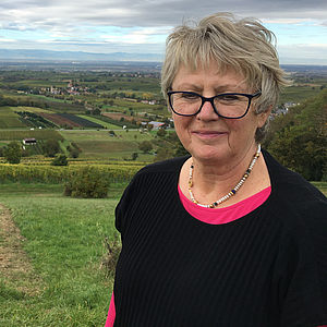 Porträt einer älteren Frau mit kurzen grauen Haaren und schwarzer Brille, trägt Perlenkette, bekleidet mit schwarzem Pullover und pinkem Untershirt, vor weiter Hügellandschaft mit Weinbergen und Feldern.
