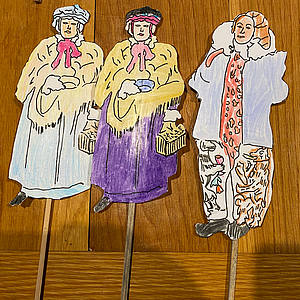 Drei handkolorierte Papierfiguren auf Holzstäben in historischen Kostümen: zwei Frauen mit Schultertüchern und kleinen Körben, eine männliche Figur mit Rüschenkragen und gemusterten Hosen, auf Holzuntergrund.