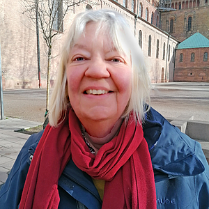 Porträt einer lächelnden älteren Frau mit weißem Haar, rotem Schal und blauer Jacke vor der Backsteinfassade eines historischen Kirchengebäudes.