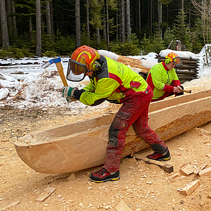 Zwei Handwerker in Schutzkleidung und Helmen bearbeiten im Wald einen ausgehöhlten Baumstamm mit Axt und Stechbeitel zu einem Kanu, Holzhackschnitzel und Schnee am Boden.
