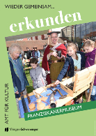 Vor dem Franziskanermuseum zeigt eine erwachsene Person in historischer Kleidung Kindern handgefertigte Holzobjekte auf einem Tisch; mehrere Kinder schauen neugierig.