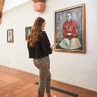 Eine Besucherin steht in einem Museum vor einem großen, gerahmten Porträt einer sitzenden Person in roter Kleidung an einer weißen Wand; weitere kleine Bilder und ein gefliester Boden sind sichtbar.