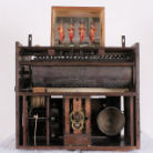Holzgehäuse eines mechanischen Musikinstruments: sichtbare gezapfte Walze, Zahnräder und Hebel, rechts eine große Messingglocke; oben drei bemalte Holzfiguren in kleinem Kasten.