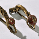Zwei goldene spiralförmige Schmuckbeschläge mit rot emaillierten Rundintarsien, leicht patiniert, auf hellem Hintergrund.