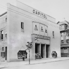 Schwarzweißfoto eines Kinogebäudes mit großer Aufschrift CAPITOL über dem Eingang, markante Fassadenornamente, zwei Türen unter einem kleinen Vordach mit Marquee und Filmplakaten, nebeneinanderstehende Schaukästen, angrenzende Wohnhäuser
