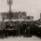 Historische Schwarzweißaufnahme: Gruppe von Männern in Uniform und Zivil vor Stadthäusern, vorn Kapelle mit Trommeln und Blasinstrumenten, im Hintergrund dreistöckiges Gebäude und Sendemast.