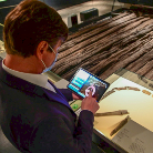 Eine Person mit Mund-Nasen-Schutz sitzt an einem Tisch, hält ein geöffnetes Buch und bedient ein Tablet; ein geöffnetes Notizbuch liegt daneben.