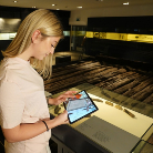 Besucherin hält ein Tablet und betrachtet hinter einer Glasvitrine beleuchtete Exponate in einer Ausstellung des Franziskanermuseums.