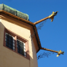 Eckansicht einer hellen historischen Hausfassade mit zwei vergoldeten Wasserspeiern als Dachrinnen-Ausläufe, die seitlich hervorstehen; darunter zwei kleine Fenster mit Gittern; klarer blauer Himmel im Hintergrund.