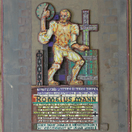 Vergoldetes Relief an einer Wand: kniender Ritter in Rüstung, rechte Hand hebt ein rundes Attribut, linke stützt ein hohes Kreuz; darunter buntes Wappen und mehrzeilige Inschriftstafel.