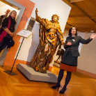 Museumsszene: Frau erläutert eine große, vergoldete Heiligenstatue in reich drapierten Gewändern mit erhobenem Arm, stehend auf einem Sockel; Ausstellungsschild und warme Raumbeleuchtung