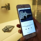 Hand hält Smartphone, auf dessen Bildschirm eine Nahaufnahme eines verzierten historischen Metallobjekts mit Begleittext zu sehen ist; im Hintergrund steht dasselbe Objekt in einer Vitrine.