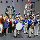 Auf der Bühne: Gruppe in historischen blauen Uniformen mit weißen Hosen und Federhüten, marchierende Musiker mit Trommel und Blasinstrumenten; links zwei Personen in großen Pferdekostümen tragen eine dekorative Girlande.