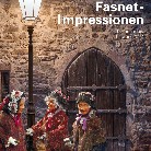 Drei vermummte Fasnetsfiguren in traditionellen Kostümen und Holzmasken stehen abends vor einer steinernen Mauer mit rundem Holzportal; eine Laterne beleuchtet den verschneiten Platz.