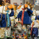 Gruppe traditioneller Maskenfiguren: mehrere Gestalten in blauen Mänteln, Pelzmützen und orangefarbener Haarpracht halten lange Stangen; vorne eine geschmückte Tiermaske mit bunten Papierstreifen.