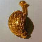 Goldener, filigran verzierter eiförmiger Anhänger mit Öse und reich ornamentiertem Netz- und Punktmuster, liegt schräg auf hellem Untergrund und wirft Schatten.
