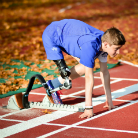 Sportler in blauer Laufkleidung mit Unterschenkelprothese in Startblockposition auf roter Tartanbahn, bereit zum Sprint.