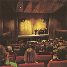 Theatersaal von hinten: Zuschauer in roten Sitzen sehen auf eine beleuchtete Bühne mit Vorhang, auf der drei Personen stehen und sprechen.