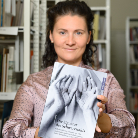 Lächelnde Person hält frontal ein Buch mit einem Cover, das ineinandergreifende Hände zeigt; im Hintergrund sind Bücherregale sichtbar.
