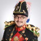 Porträt eines lächelnden Mannes in historischer Festuniform: schwarzer Dreispitz mit bunten Federn, runde Brille, Schnurrbart und Kinnbart, goldverziertes schwarzes Jackett mit roten Details und Orden.