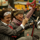 Zwei verkleidete Erwachsene in nostalgischer Kleidung auf einem kleinen offenen Wagen; vorne eine Frau mit Schirmmütze, dahinter ein Mann mit Schutzbrille; beide lachen und winken, Umzugspublikum im verschwommenen Hintergrund.