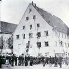 Historische Schwarzweißaufnahme: großes spitzgiebeliges Haus mit mehreren Fensterreihen; eine Menschenmenge steht davor, einige Personen auf langen Leitern an der Fassade.