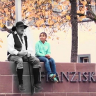 Skulptur eines sitzenden Mannes mit Hut und Hosenträgern auf einer Mauer, daneben ein in türkis gekleidetes Kind; im Hintergrund Herbstbäume und an der Mauer der Schriftzug FRANZIS