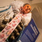 Ausstellungsobjekt: geschnitzter Kopf mit bemaltem Gesicht, angehängt an ein Stoffband mit rosafarbenem Blumenmuster; darunter zahlreiche kleine Metallglocken in Kettenform; Museumsbeschriftung im Hintergrund.