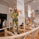 Museumsausstellung: Mehrere bemalte, flache Holzfiguren in farbenfrohen, mittelalterlich anmutenden Kostümen und Tierdarstellungen auf einfachen Holzgestellen in einem hellen Ausstellungsraum