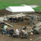 Archäologische Ausgrabung: mehrere Personen graben in einem rechteckigen Schacht unter einem weißen Sonnendach, mit Eimern, Werkzeugen und einer Schubkarre auf freiem Feld.