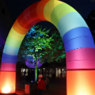 Beleuchtete, regenbogenfarbene Bogeninstallation in einem nächtlichen Innenhof vor dem Franziskanermuseum; zwei Personen stehen unter dem Bogen, im Hintergrund ein grün-blau beleuchteter Baum und historische Gebäude.
