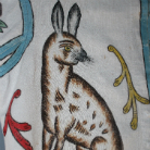 Stilisierter braun gefleckter Hase mit aufgerichteten Ohren auf hellem Stoff, umgeben von dekorativen Ranken in Rot, Blau und Gelb.
