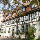Außenansicht des Franziskanermuseums: mehrgeschossiges Fachwerkhaus mit dunklen Holzbalken, zahlreichen Fenstern, dekorativem Eingang, Satteldach und begrünten Pflanzen; Vordergrund mit herbstlichen Baumzweigen.