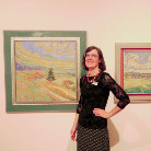 Im Franziskanermuseum Villingen-Schwenningen steht eine Frau lächelnd vor zwei gerahmten Landschaftsgemälden an einer weißen Ausstellungswand; die Bilder zeigen Hügel, Bäume und pastellfarbene Himmel.