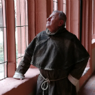 Lebensgroße Figur eines Franziskanerbruders in braunem Habit mit Kordel, stehend am gotischen Fenster, mit erhobenem Blick nach draußen, sachte vom Licht illuminiert, vermittelt kontemplative Atmosphäre.