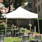 Weißes Pavillonzelt mit Tisch und stehender Person, davor Reihen leerer Stühle, zwei Blumenarrangements auf Säulen und einige Luftballons im grünen Innenhof des Franziskanermuseums vor historischen Gebäudefassaden.