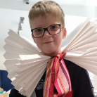 Porträt eines Jungen mit Brille, der eine große weiße, gefaltete Rüschenkragen‑Attrappe aus Papier trägt, vorne mit einem rot‑gestreiften Band gebunden; leicht lächelnd.