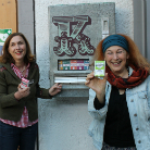 Zwei lächelnde Frauen vor einer Hauswand zeigen auf eine wandmontierte, verzierte Metall-Einwurfbox des Franziskanermuseums; eine von ihnen hält eine kleine Karte oder einen Flyer.