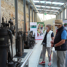Zwei Museumsbesucher, eine Person mit Rollator und ein Mann mit Hut, betrachten in einem verglasten Gang nebeneinander stehende historische Dampfmaschinen und Informationstafeln im Franziskanermuseum Villingen-Schwenningen.