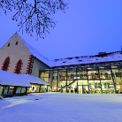 Verschneiter Innenhof des Franziskanermuseums bei Dämmerung, links historische Kirchenfassade mit rot gerahmten Spitzbogenfenstern, rechts moderne verglaste Eingangsfront mit beleuchtetem Inneren