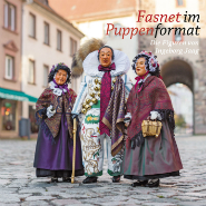 Vier traditionell kostümierte Fasnetsfiguren in bunten Röcken, bestickten Schürzen und gestrickten Kopfbedeckungen stehen auf Kopfsteinpflaster vor Häuserfassaden; zwei tragen geflochtene Körbe.