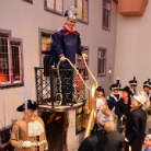 Große lebensgroße Figur in blauer Uniform auf einem erhöhten Balkon, darunter Menschen in historischen Uniformen und Trachten bei einer Straßenszene.