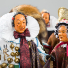 Zwei traditionell kostümierte Figuren mit geschnitzten Holzmasken, großen weißen Rüschenkragen, bunten Gewändern und angebrachten Glocken; im Hintergrund eine weitere ähnliche Gestalt.