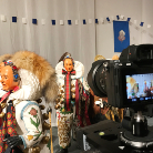 Zwei fastnachtliche bemalte Holzmasken mit aufwändigen Kostümen (Pelzkrägen, karierte Stoffe) stehen in einem Ausstellungsraum des Franziskanermuseums vor einer Kamera, die sie in Nahaufnahme aufzeichnet.